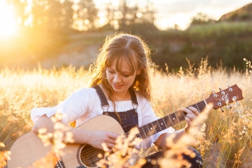 Young woman playing guitar in grass backlit by golden light - Australian Stock Image