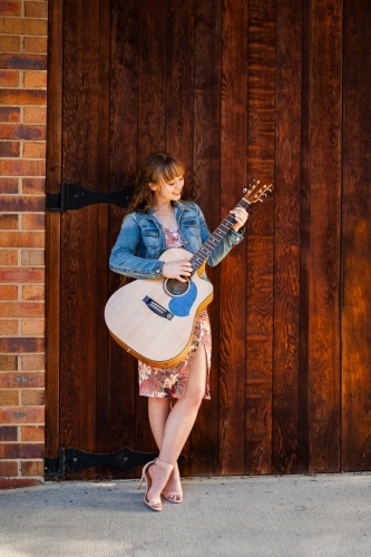 Young woman playing guitar outside building with wooden door - Australian Stock Image