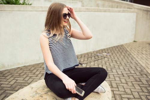 Young woman sitting against a wall with a phone and sunglasses