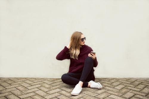 Young woman sitting against a wall with a phone and sunglasses - Australian Stock Image