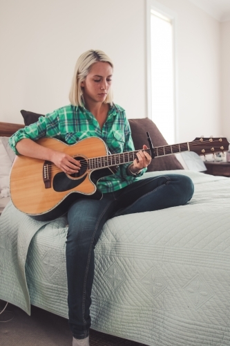Young woman sitting on a bed looking down as she plays the guitar - Australian Stock Image