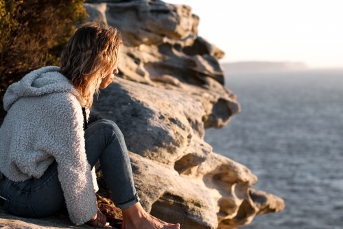 Young woman sitting on a clifftop looking out at the ocean at sunrise - Australian Stock Image