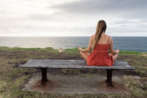 Young woman sitting on a park bench looking out to sea in a meditation pose
