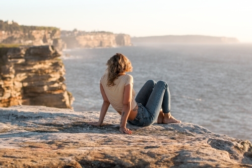 Young woman sitting on coastal clifftop at sunrise looking out to sea - Australian Stock Image
