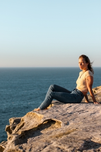 Young woman sitting on coastal clifftop at sunrise looking out to sea - Australian Stock Image