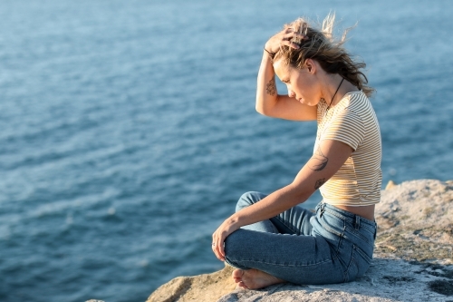 Young woman sitting on coastal clifftop at sunrise looking out to sea - Australian Stock Image
