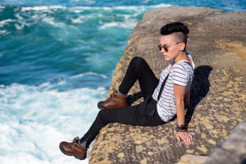 Young woman sitting on the edge of a cliff by the ocean - Australian Stock Image