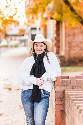Young woman standing on street wearing hat and scarf smiling leaning on brick wall - Australian Stock Image
