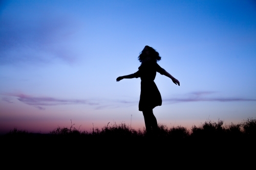 Young woman twirling silhouetted against pastel dusk sky - Australian Stock Image