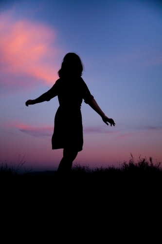 Young woman twirling silhouetted against pastel dusk sky - Australian Stock Image