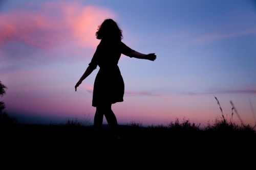 Young woman twirling silhouetted against pastel dusk sky - Australian Stock Image