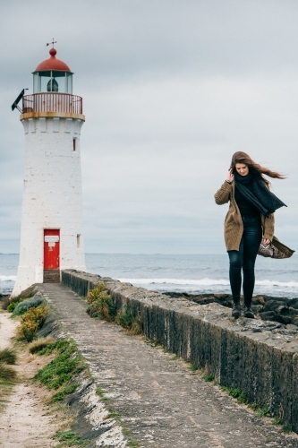 Young woman walks along the wall in front of lighthouse. - Australian Stock Image