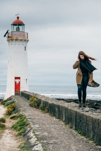 Young woman walks along the wall in front of lighthouse.