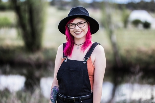 Young woman with pink hair glasses and hat smiling standing near river water - Australian Stock Image