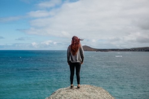 Young woman with red hair standing on rock looking at ocean - Australian Stock Image