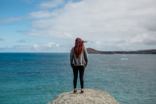 Young woman with red hair standing on rock looking at ocean