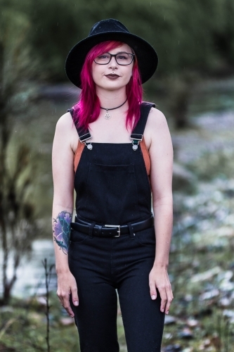 Young woman with tattoo pink hair hat and glasses standing near river - Australian Stock Image