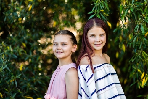 Younger and older tween sisters stand back to back smiling